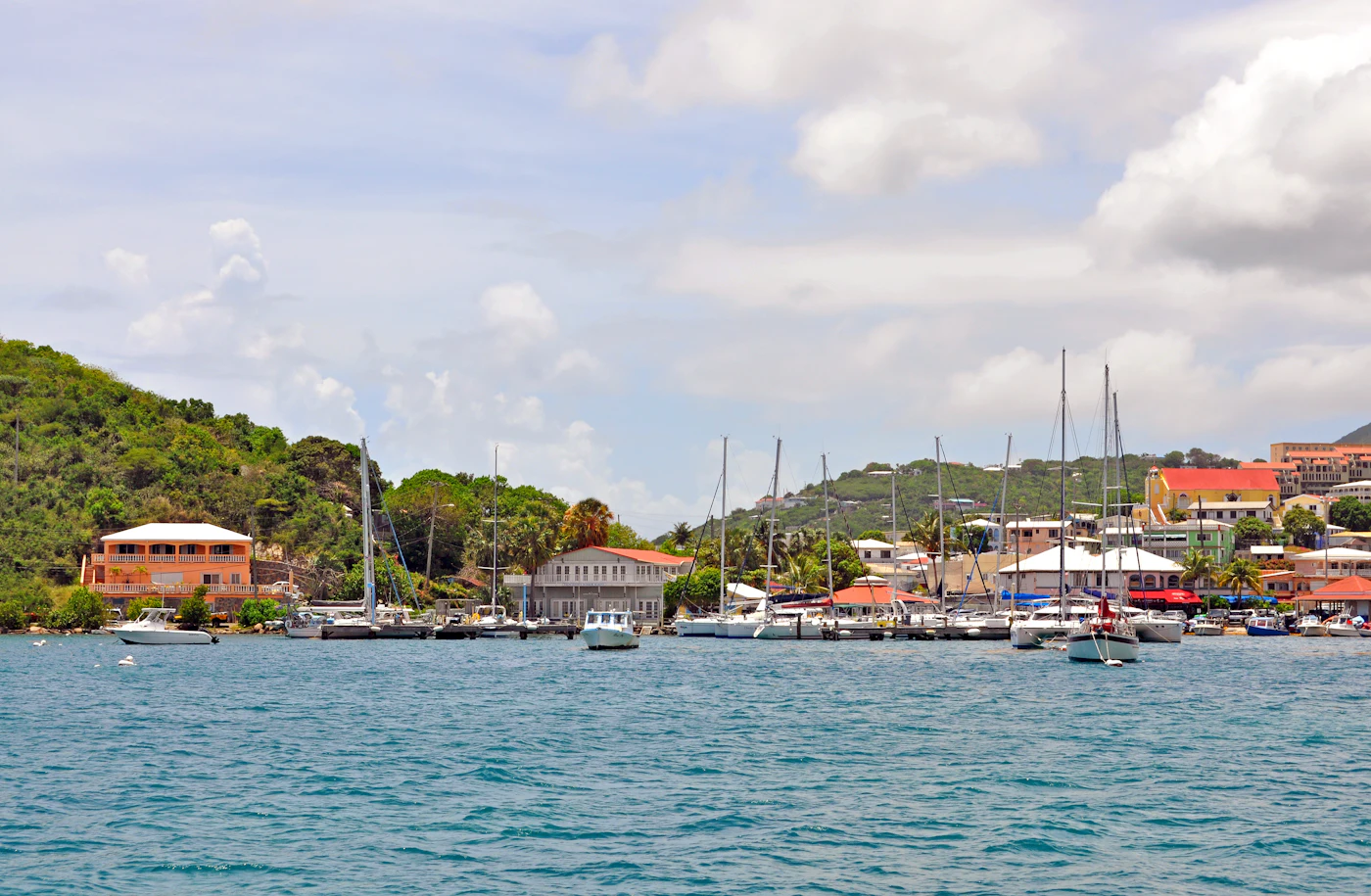 Sailboats and harbor buildings along a calm Caribbean bay with green hills on a bright day